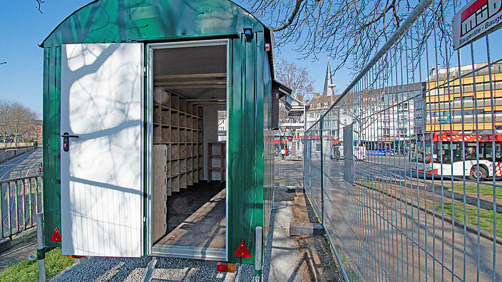 Ein ausgebauter, grün gestrichener Bauwagen dient als Taubenhaus am Zentralen Busbahnhof in Bonn. Neben dem Bauwagen steht ein Bauzahn, der das Taubenhaus vor Vandalismus schützt.