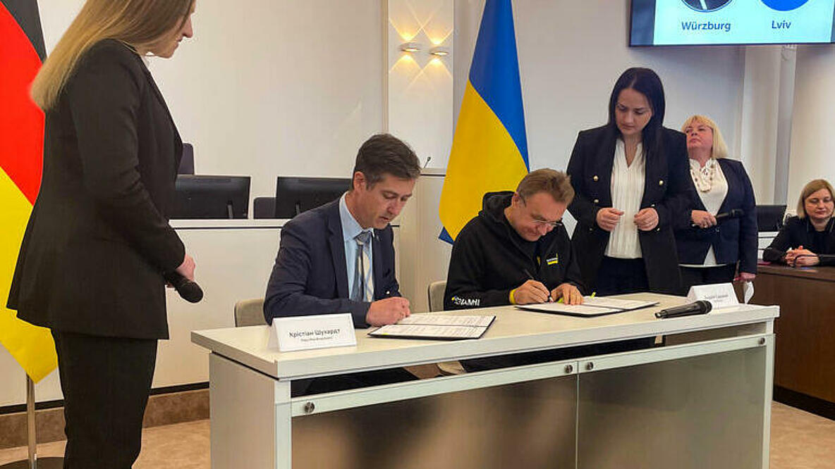 Zwei Männer unterschreiben eine Urkunde mit den deutschen und ukrainischen Flaggen im Hintergrund